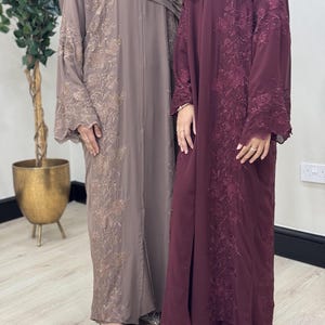 May include: Two floor-length abayas, one in a muted taupe and the other in deep burgundy. Both feature elaborate floral embroidery and matching head coverings. The abayas have long sleeves, perfect for special occasions.