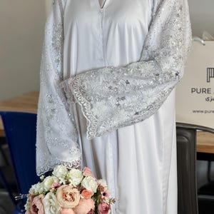 May include: White abaya with detailed silver embroidery on the sleeves. The sleeves have floral and sequin embellishments. The abaya is worn with a white head covering and a bouquet of pink and white flowers.