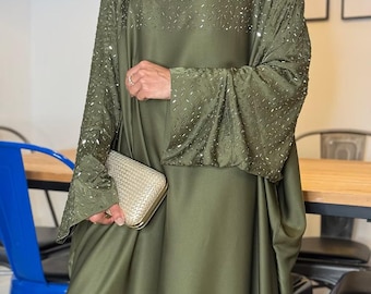 May include: Olive green abaya with embellished sleeves and bodice. The flowing dress features a matching head covering and is paired with a gold clutch. The abaya is a modest garment, suitable for special occasions.