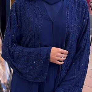 Könnte beinhalten: Eine marineblaue Abaya mit aufwendigen Perlenarbeiten an den Ärmeln und Vorderteilen. Das Kleidungsstück hat ein fließendes Design und einen bescheidenen Ausschnitt. Die Ärmel sind lang und ausgestellt, und der Stoff hat einen subtilen Glanz.