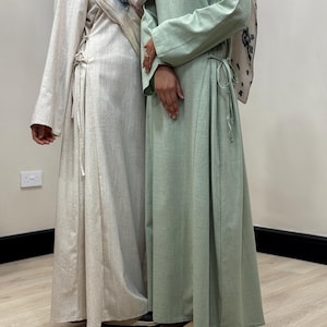 May include: Two long-sleeved maxi dresses, one in beige and one in sage green. Both garments feature a side-tie closure at the waist with thin fabric strings, a round neckline, and a floor-length skirt. The dresses are worn with headscarves.