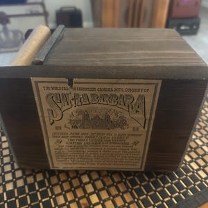 May include: A dark brown wooden pinhole camera with a label that reads "Santa Barbara." The camera has a rectangular shape with a roll on top. The label includes text about the camera's features and accessories.