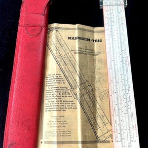 May include: Vintage Mannheim-Trig slide rule with a red leather case and instruction manual. The slide rule has a white and silver frame with black markings. The manual is tan with black text and diagrams.