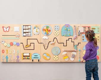 Könnte beinhalten: Eine Holz-Aktivitätstafel mit verschiedenen interaktiven Elementen, darunter ein Schmetterling, ein Lichtschalter, eine Uhr, Zahnräder und eine Jacke. Die Tafel ist an einer grauen Wand befestigt, und ein Kind interagiert damit.