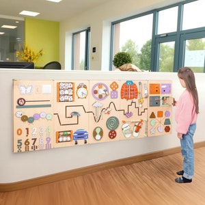 Op de afbeelding: Een houten activiteitenwand met verschillende interactieve elementen, waaronder een klok, een vlinder, een doolhof, een auto en een getallenlijn. De wand is gemonteerd op een witte muur in een kamer met een lichte houten vloer. Een jong meisje staat naast de muur en interageert met het activiteitenpaneel.