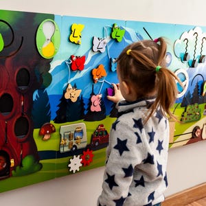 Puede incluir: Una colorida pared de actividades de madera con varios elementos interactivos, que incluyen un árbol con una puerta corredera, una autocaravana, un sistema de engranajes y una actividad de lazado de mariposas. Un niño está interactuando con la pared, demostrando sus características educativas y atractivas.