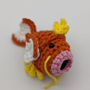 May include: A crocheted orange, pink, white, and yellow fish with black eyes. The fish has a yellow tail fin and a pink mouth.