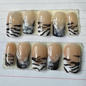 May include: A set of ten artificial nails with a beige base, black tiger stripe design, and a touch of silver glitter. The nails are oval-shaped and arranged in two rows on a clear adhesive strip.