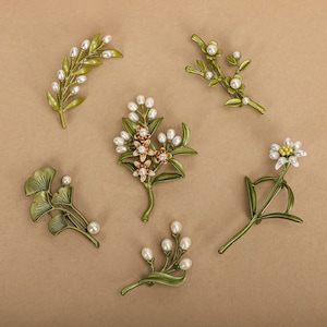 May include: A collection of floral brooches in different designs. Each brooch has a green enamel base with pearl accents. Styles include leaf, branch, and flower shapes. The brooches are displayed on a neutral tan background.