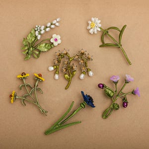 May include: An assortment of floral brooches featuring various flower designs, including daisies, forget-me-nots, and a cluster of small white flowers. The brooches are made of metal and have a vintage aesthetic.