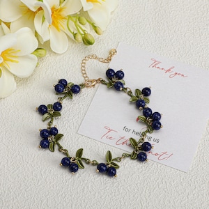 May include: A gold-coloured bracelet with dark blue bead clusters, like blueberries, and green leaf details. The bracelet is on a white surface with a card that says "Thank you".