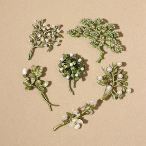 Könnte beinhalten: Eine Sammlung von sechs grünen Metallbroschen in Form verschiedener botanischer Elemente wie Zweige, Blätter und Blüten. Jedes Stück ist mit kleinen weißen Kunstperlen verziert, die wie Knospen oder Blüten an den Stielen angeordnet sind.