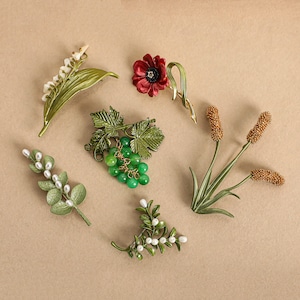 Puede incluir: Un surtido de broches florales y botánicos. La colección incluye una amapola roja, un racimo de uvas verdes y ramitas de hojas con cuentas blancas. Los broches son en tonos verdes, rojos y dorados, sobre un fondo neutro.