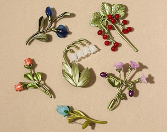 May include: A collection of six floral and botanical brooches featuring green metal stems and leaves. The set includes blue buds, red berries, white lily of the valley bells, orange rosebuds, purple and pink flowers, and a single light blue bud.