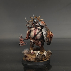 Puede incluir: Una figura en miniatura pintada de un guerrero corpulento con armadura oxidada y un casco con cuernos. La figura sostiene un hacha de batalla y un escudo circular, de pie sobre una base rocosa decorada con pequeñas calaveras y hierba.