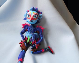 Puede incluir: Una muñeca artesanal colorida con tela azul, verde y rosa, una cara pintada y detalles de cuentas. La muñeca está sentada sobre un fondo de tela azul claro.