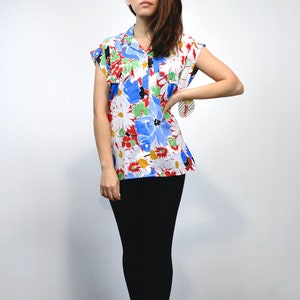 May include: A short-sleeved, button-down shirt with a vibrant floral pattern. The shirt features a white background with large blue, red, and yellow flowers. The collar and sleeves are trimmed in white. The model is wearing black pants.