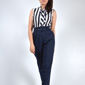 May include: A woman wearing a white and black striped sleeveless blouse with a bow tie and a navy blue high-waisted pair of pants with a belt.