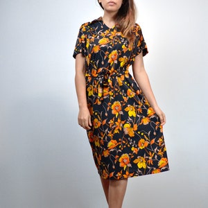 Puede incluir: Un vestido midi negro con estampado floral con flores naranjas y amarillas. El vestido tiene manga corta y un lazo en la cintura.