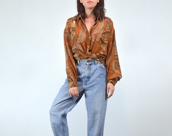 May include: A brown button-down shirt with a paisley pattern in shades of gold, red, and green is knotted at the waist. The shirt is paired with light blue denim jeans.