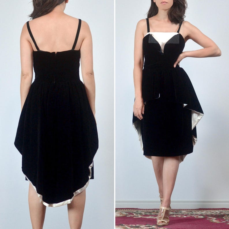 May include: A black velvet dress featuring a high-low hemline with a white satin lining, thin shoulder straps, and a structured bodice with a white satin insert at the neckline. The garment displays a fitted waist and a flared skirt silhouette.