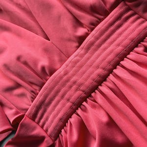 May include: Close-up of a coral fabric with a pleated design. The fabric has a smooth, silky texture with a central panel featuring vertical stitched lines. The overall aesthetic suggests a garment or textile.