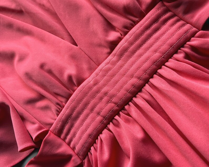 May include: Close-up of a coral fabric with a pleated design. The fabric has a smooth, silky texture with a central panel featuring vertical stitched lines. The overall aesthetic suggests a garment or textile.