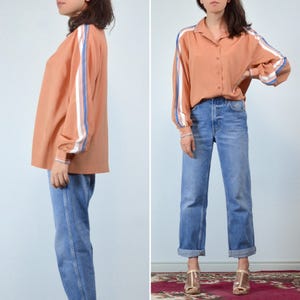 May include: A peach-colored button-down shirt with blue and white stripes on the sleeves, paired with light blue denim jeans. The shirt has a collared neckline and long sleeves. The jeans are cuffed at the ankles.