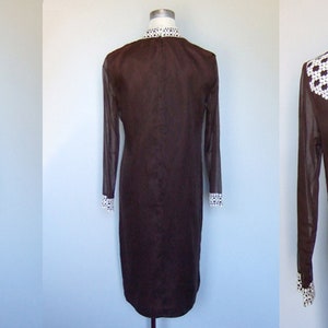 May include: A brown dress with a white, geometric patterned collar and cuffs. The dress has long, sheer sleeves and a straight silhouette. The dress is displayed on a mannequin, showcasing its vintage style and design.