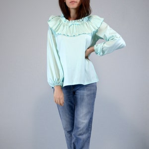 Pode incluir: Blusa azul clara com gola de babados plissados e mangas compridas. A blusa tem decote e punhos franzidos. A modelo est&aacute; usando jeans azuis. O fundo &eacute; cinza claro liso.