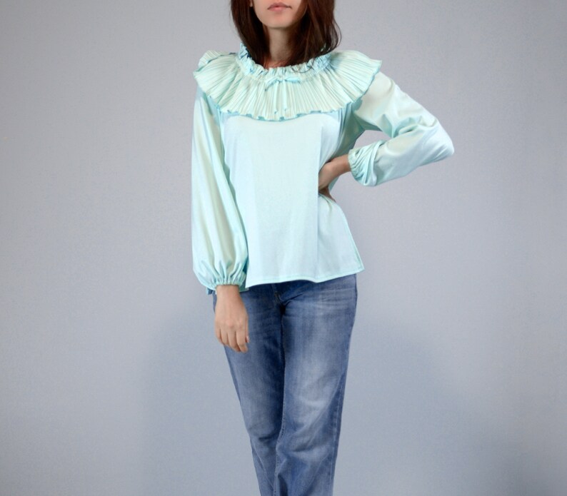 Pode incluir: Blusa azul clara com gola de babados plissados e mangas compridas. A blusa tem decote e punhos franzidos. A modelo est&aacute; usando jeans azuis. O fundo &eacute; cinza claro liso.
