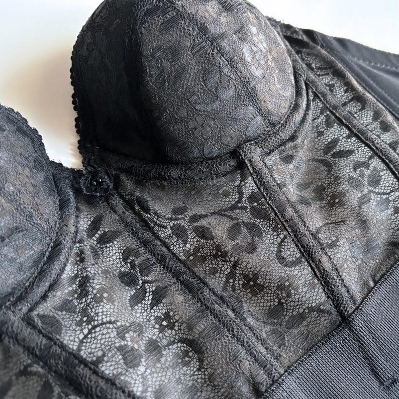 May include: Close-up of a black corset with intricate floral lace overlay. The corset features a structured bodice with molded cups and a smooth, solid fabric on the sides and back. The lace detailing adds a delicate texture to the garment.