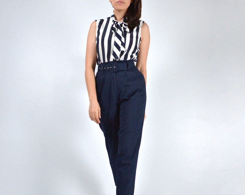 May include: A woman wearing a white and black striped sleeveless blouse with a bow tie and a navy blue high-waisted pair of pants with a belt.