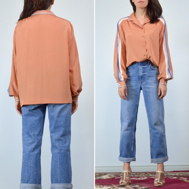 May include: A peach-colored button-down shirt with white and blue striped accents on the sleeves and collar. The shirt is paired with blue jeans. The model is wearing gold-colored heels.