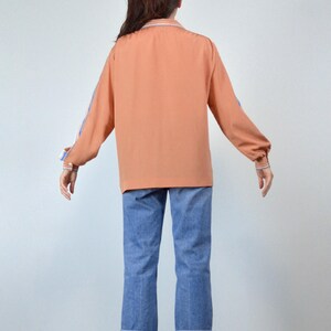 May include: A peach-colored long-sleeve shirt with blue and white stripes on the sleeves and cuffs. The shirt is paired with blue denim jeans. The outfit is viewed from the back, showcasing the details of the clothing.