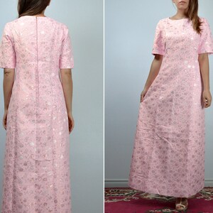 May include: A light pink maxi dress with short sleeves and a round neckline. The dress features a floral pattern in silver and white. The dress is floor-length and has a simple, elegant design, suitable for formal occasions.