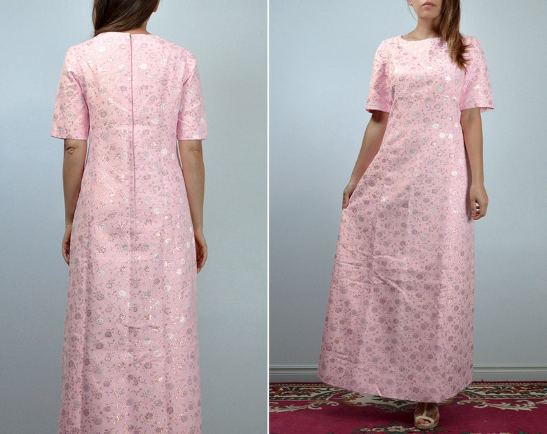 May include: A light pink maxi dress with short sleeves and a round neckline. The dress features a floral pattern in silver and white. The dress is floor-length and has a simple, elegant design, suitable for formal occasions.