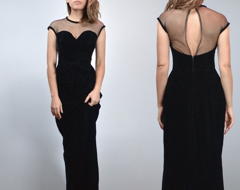 May include: A black velvet evening gown with a sheer neckline and cap sleeves. The bodice features a sweetheart neckline and a fitted waist. The back of the dress has a sheer panel. The dress is floor-length.