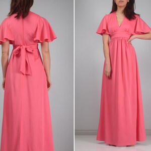 May include: A coral maxi dress with short, flared sleeves and a V-neck. The dress has a fitted waist and a flowing skirt. The back of the dress features a tie detail at the waist. The dress is made of a smooth, solid fabric.
