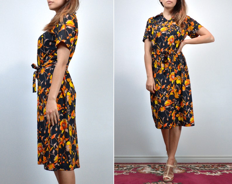 Puede incluir: Un vestido midi negro con estampado floral en naranja y amarillo. El vestido tiene un lazo en la cintura y mangas cortas.