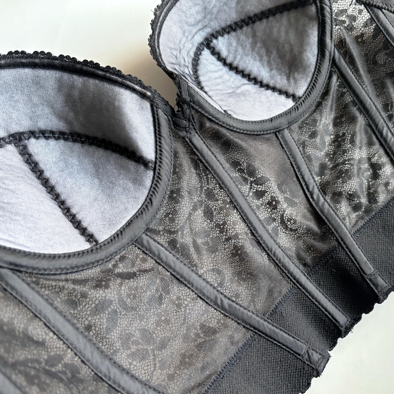 May include: A close-up of a black corset with intricate lace detailing. The corset features structured boning, a scalloped trim, and a satin finish. The interior lining is a light gray color, providing a contrast to the dark exterior. The overall design is elegant and sophisticated.
