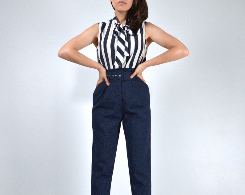 May include: A woman wearing a navy blue and white striped sleeveless blouse with a bow tie and a pair of high-waisted navy blue pants with a belt.