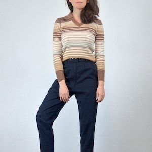 May include: A vintage-style collared sweater with horizontal stripes in shades of brown, beige, and cream. The long-sleeved top is paired with high-waisted, dark navy blue trousers. The outfit is a classic retro look.