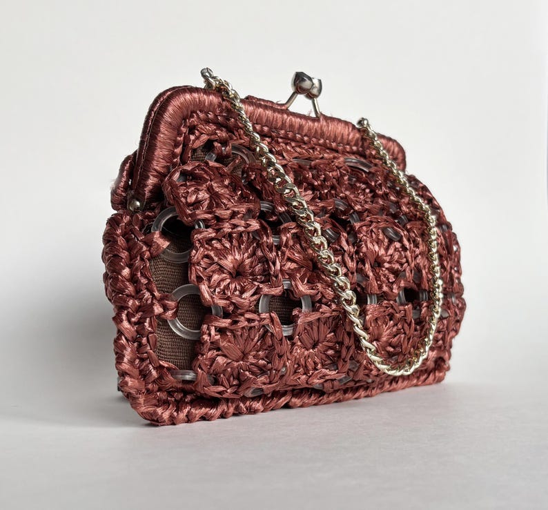 May include: A reddish-brown, crocheted handbag with a silver chain strap. The bag features a kiss-lock closure and a unique design incorporating metal ring accents. The texture of the bag is intricate and detailed, showcasing a vintage aesthetic.