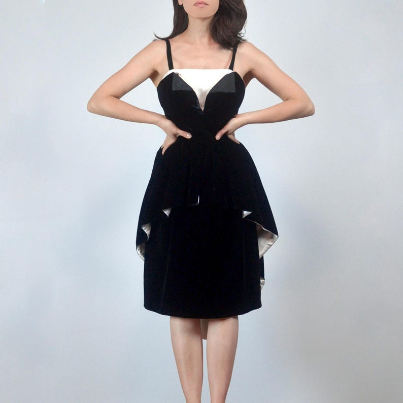 May include: A knee-length black velvet dress featuring a white bodice panel, thin shoulder straps, and a layered peplum skirt design. The garment has a structured silhouette with a contrasting white lining visible beneath the black velvet fabric.