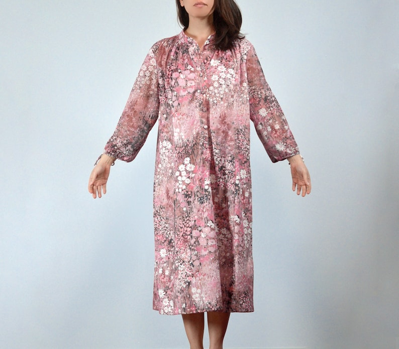May include: A vintage floral print dress in shades of pink, white, and gray. The dress features long sleeves, a mandarin collar, and a button-down front. The fabric appears to be sheer, with a delicate, flowing silhouette.