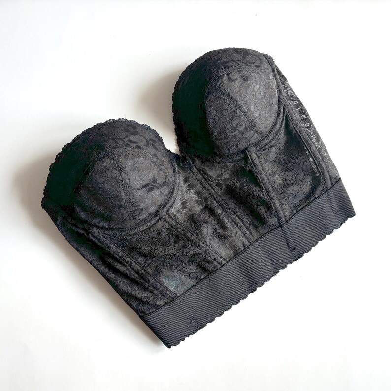 May include: Black bustier with floral lace detailing. The underbust band is a solid black with a scalloped edge. The cups are padded and the bustier has a structured silhouette. The fabric has a subtle sheen.
