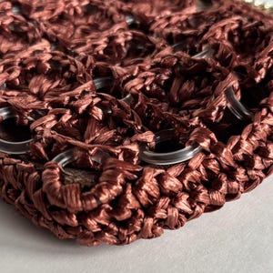 May include: Close-up of a woven, reddish-brown handbag. The bag features a textured, open weave design with clear, circular eyelets. The material appears to be a raffia-like fiber, creating a natural, earthy aesthetic.