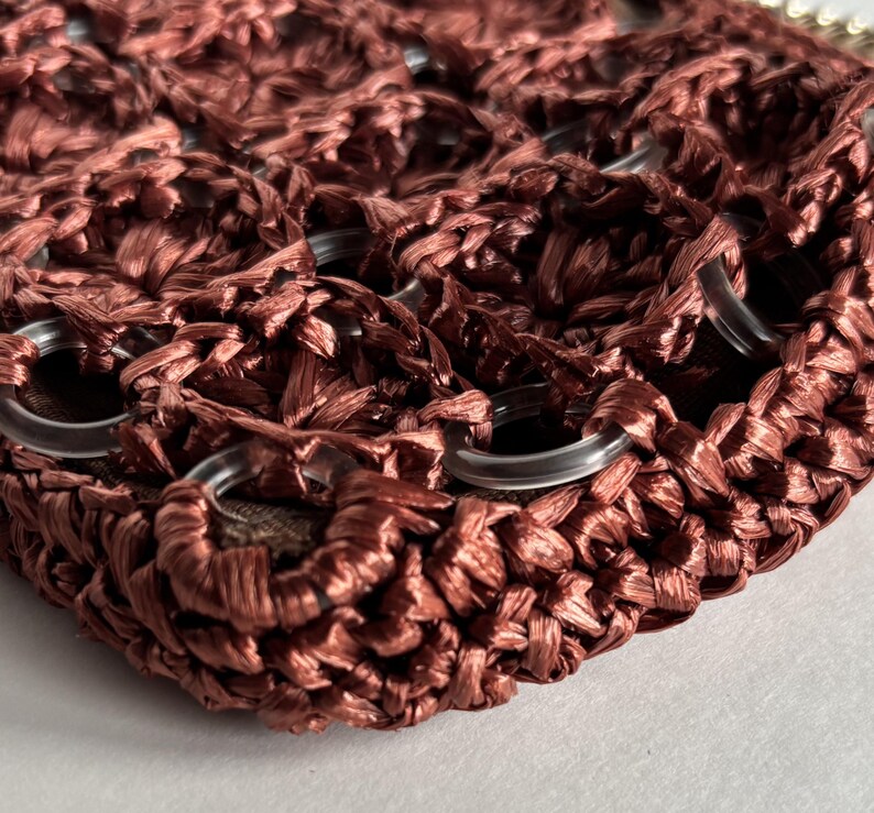 May include: Close-up of a woven, reddish-brown handbag. The bag features a textured, open weave design with clear, circular eyelets. The material appears to be a raffia-like fiber, creating a natural, earthy aesthetic.