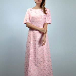 May include: A light pink, floor-length dress with short sleeves. The dress features a subtle floral pattern in a lighter shade of pink and silver. The dress has a boat neckline and a simple, elegant design.